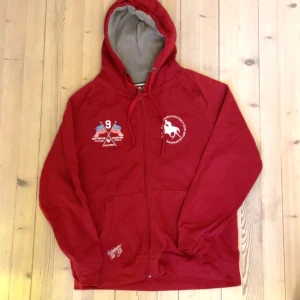 Röd hoodie från Huntington Polo Club - Säljer en röd hoodie från Huntington Polo Club med dragkedja och tryck på bröstet. Den har en justerbar huva och två fickor framtill. Perfekt för en avslappnad stil.