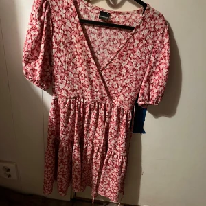 Somrig klänning med blommor  - Söt blommig klänning från New Look i rött och vitt. Den har en v-ringad design med korta ärmar och en luftig kjol. Perfekt för sommarens alla tillfällen!