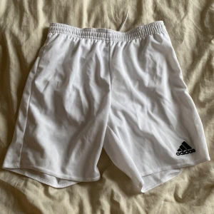 Vita träningsshorts från Adidas - Säljer ett par vita träningsshorts från Adidas. De har en elastisk midja med snörning för justerbar passform. Perfekta för sport och aktivitet. Adidas-loggan är tryckt på benet. Storlek 11-12 år, skulle säga runt om strl 146💗