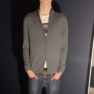 Grå kofta med dragkedja - Säljer en stilren grå kofta i merino ull med dragkedja. Perfekt för kyligare dagar och enkel att matcha med olika outfits. Koftan har långa ärmar och ribbade muddar för en bekväm passform. Modellen på bilden är 187 65kg och tröjan sitter bra på dem.