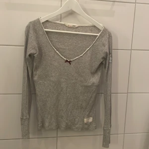 Grå ribbad tröja från Odd Molly - Säljer en grå ribbad tröja från Odd Molly med u-ringning och spetsdetaljer. Tröjan har en liten rosett framtill och ett märke på ärmen med texten 'Lost on Purpose'. 
