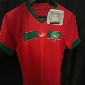 Marocko fotbollströja från Puma Stl Xs  - !!!!!!!!LÄS!!!!!                                                           Snygg röd fotbollströja från Puma med gröna detaljer och korta ärmar. Tröjan har ett emblem på bröstet och Puma-loggan. Perfekt för fotbollsfans som vill visa sitt stöd. Helt ny! Har bara testat den dock har jag tagit av lapparna nyligen för jag tänkte börja anvönda den men fortfarande inte anvönd😂 lapparna kan man få med i vilket fall ☺️👍🏽