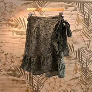 Svartprickig omlottkjol med volang - Charmig omlottkjol med svart prickigt mönster och volangdetaljer. Kjolen har en knytning i sidan som ger en fin passform. Perfekt för en stilfull look.