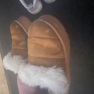 Bruna tofflor med fuskpäls - Mysiga bruna tofflor med vit fuskpäls runt kanten. Perfekta för att hålla fötterna varma under kalla dagar. Tofflorna har en mjuk och bekväm insida.