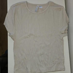 Beige ribbad t-shirt från H&M Divided - Säljer en beige ribbad t-shirt från H&M Divided. Den är kortärmad och tillverkad i en mjuk bomullsblandning med elastan för extra komfort. Perfekt för en avslappnad stil.