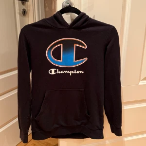 Svart hoodie från Champion - Hej! Säljer en svart hoodie från Champion med  logga i blått och svart på framsidan. Tröjan är väldigt bekväm. 