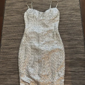 Paljett silverklänning med smala axelband - Snygg glittrig silverklänning med smala axelband och en tight passform. Klänningen har en elegant öppen ryggdetalj och är perfekt för festliga tillfällen. Den är kort och har en glittrig yta som verkligen fångar ljuset.