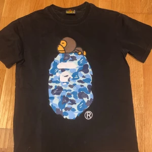 Svart t-shirt från A Bathing Ape - Säljer en svart t-shirt från A Bathing Ape med ett stort blått och vitt kamouflagemönster på framsidan. T-shirten har en kortärmad design och är tillverkad i bomull. Perfekt för dig som gillar streetwear och unika tryck. OBS 1 two 1 ‼️‼️‼️‼️Den har några små sträck i trycket men det syns inte. Det står M men passar S också. Hör av er vid funderingar!