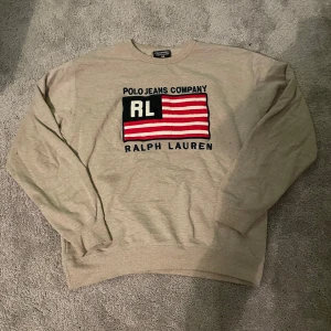 Beige tröja från Ralph Lauren - Säljer en beige tröja från Ralph Lauren med ett stort tryck av märket och en flagga i rött, svart och vitt på framsidan. Tröjan har långa ärmar och är tillverkad i bomull och polyester. Perfekt för en avslappnad stil.