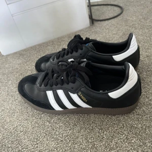 Svarta Adidas Samba sneakers - Snygga svarta Adidas Samba sneakers med vita detaljer och klassiska tre ränder på sidan. Skorna har en låg profil och är perfekta för en sportig look. De har snörning och en gummisula som ger bra grepp.