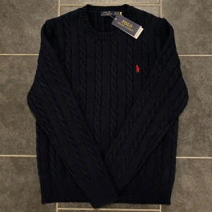 Mörkblå stickad tröja från Polo Ralph Lauren - Säljer en stilren mörkblå stickad tröja från Polo Ralph Lauren med det klassiska röda logotypen på bröstet. Tröjan har ett flätat mönster och långa ärmar, perfekt för kyligare dagar.