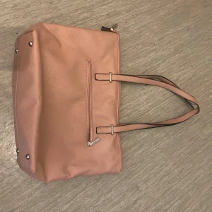 Rosa axelväska med hjärtan - Säljer en söt rosa axelväska med praktisk dragkedja och hjärtmönstrat foder. Väskan har långa handtag och en yttre ficka med dragkedja för extra förvaring. Perfekt för att bära med sig det nödvändigaste med stil. Har lite skador men påverkar inte väskan