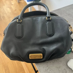 Svart handväska med gulddetaljer - Säljer en snygg svart handväska från Marc by Marc Jacobs. Väskan är i skinn och har fina gulddetaljer. Den har en praktisk dragkedja och en liten platta med logotypen på framsidan. Perfekt för både vardag och fest!