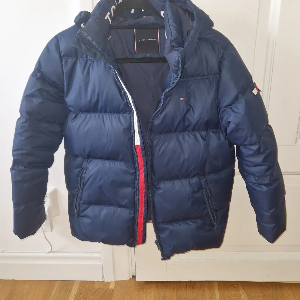 Säljer en snygg och varm mörkblå pufferjacka från Tommy Hilfiger i storlek 152. Jackan har en huva och dragkedja framtill med coola röda och vita detaljer. Perfekt för kyliga dagar!. Jackor.