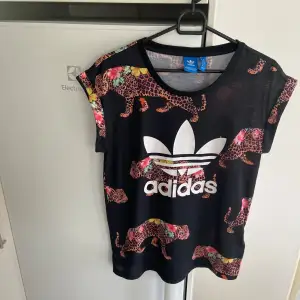 Säljer en cool svart t-shirt från Adidas i storlek M med ett färgglatt leopardmönster. T-shirten har korta ärmar och en stor vit Adidas-logga på framsidan. Nyskick kanske använd någon gång om inte enbart testad. 🐆✨går ej att köpa någonstans. 