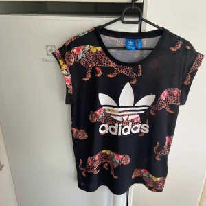 Svart t-shirt med leopardmönster från Adidas - Säljer en cool svart t-shirt från Adidas i storlek M med ett färgglatt leopardmönster. T-shirten har korta ärmar och en stor vit Adidas-logga på framsidan. Nyskick kanske använd någon gång om inte enbart testad. 🐆✨går ej att köpa någonstans. 