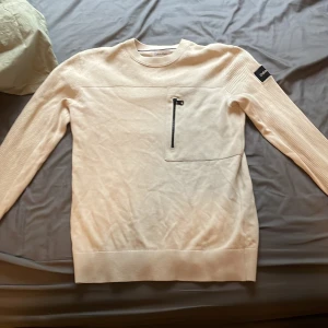 Beige tröja från Calvin Klein - Säljer en snygg beige tröja från Calvin Klein. Tröjan är ribbad på ärmarna och har en stilren design med en liten Calvin Klein-logga på ärmen. Perfekt för både vardag och lite finare tillfällen! Litet hål under högra armen 