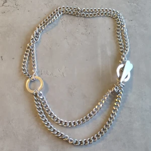 Silverfärgat halsband med dubbla kedjor - Snyggt och stilrent silverfärgat halsband med dubbla kedjor och en unik låsdesign. Halsbandet har en rund detalj med inskriptionen 'OXXO' och ett dekorativt lås som ger en modern touch. Perfekt för att lyfta vilken outfit som helst!