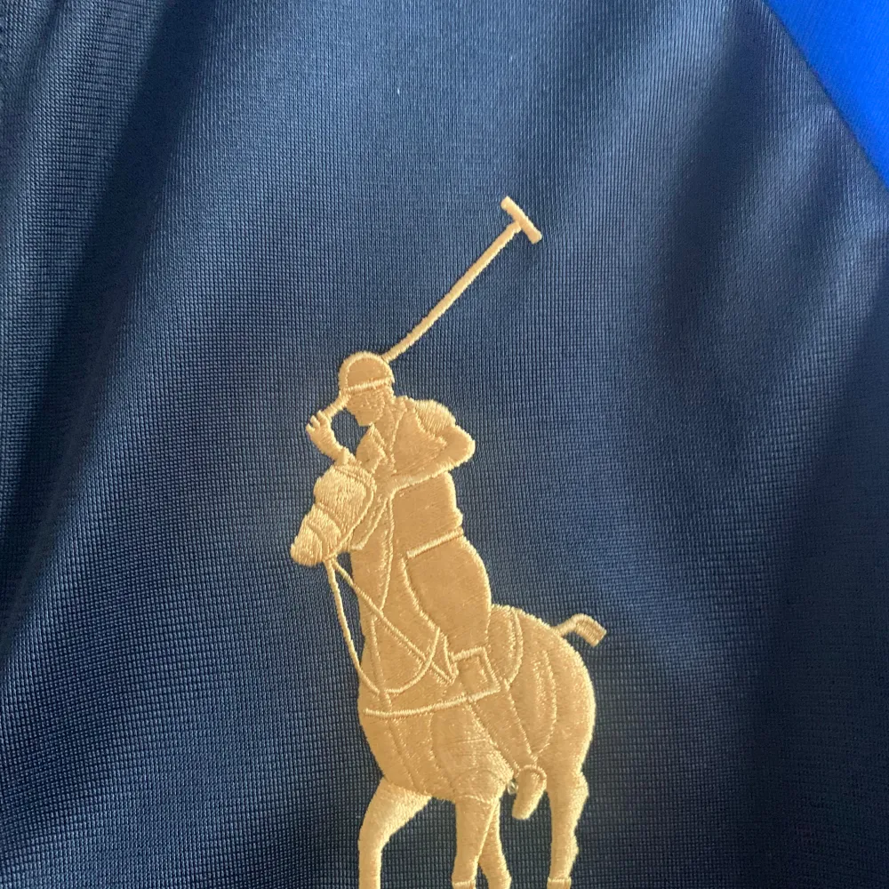 Säljer en snygg svart och blå jacka från Polo Ralph Lauren. Jackan har en dragkedja framtill och är dekorerad med broderade emblem i guld och rött på bröstet. Den har långa ärmar och är perfekt för en sportig look. Passar både till vardags och till mer avslappnade tillfällen.. Tröjor & Koftor.