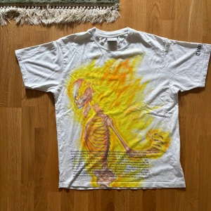 Vit t-shirt med flammande skelettmotiv från Teddy Fresh - Säljer en  vit t-shirt från Teddy Fresh med ett stort, färgglatt tryck av ett skelett omgivet av eldflammor i gult och orange. T-shirten har korta ärmar och är perfekt för en avslappnad stil. Textdetaljer finns också på framsidan