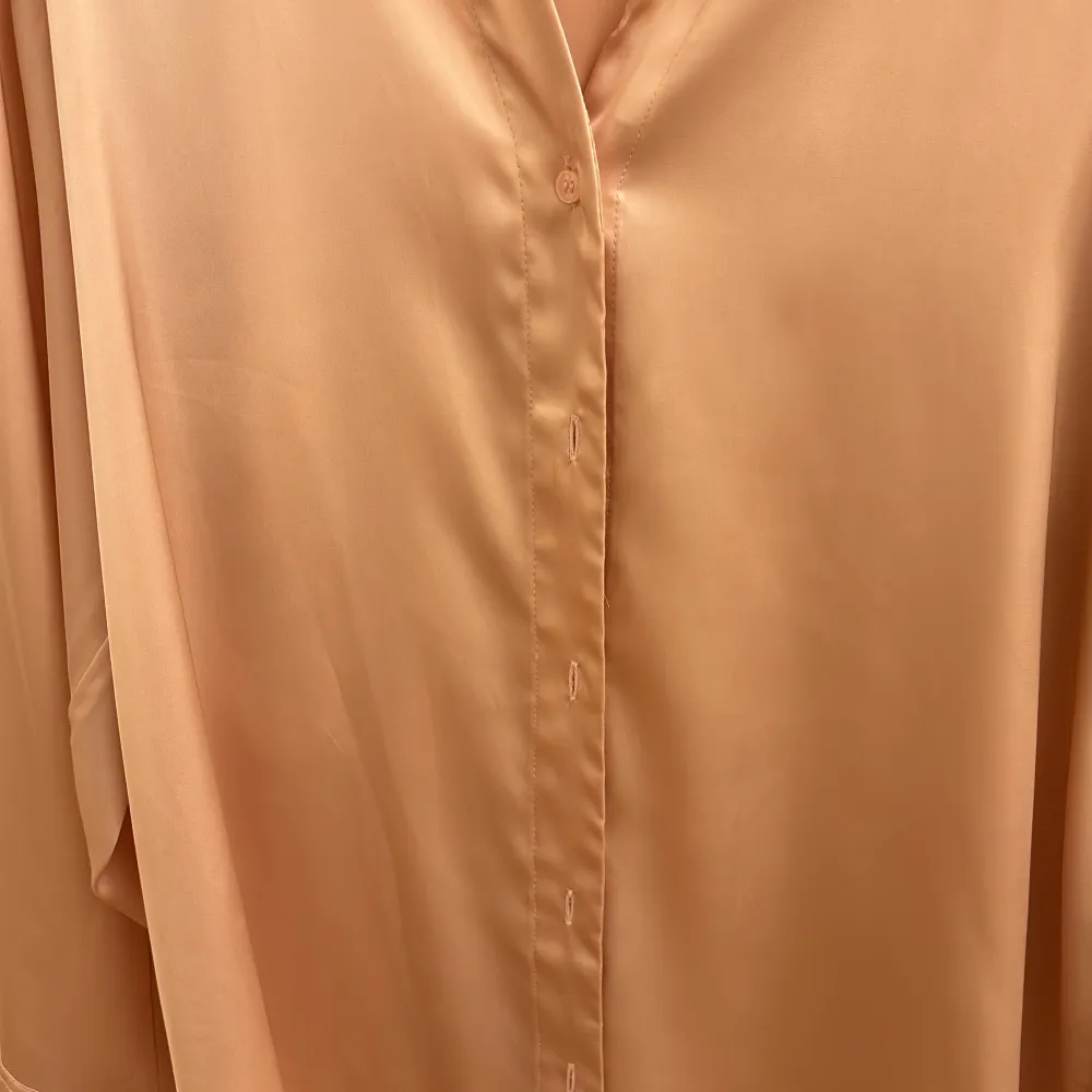 Säljer en elegant beige blus i satin från H&M. Den har en mjuk och glansig yta med långa ärmar och en klassisk krage. Blusen har en dold knäppning framtill som ger en stilren look. Perfekt för både vardag och fest!. Skjortor.