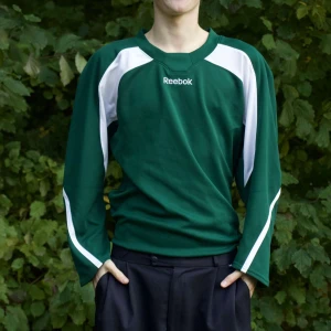 Grön långärmad tröja från Reebok - Säljer en grön långärmad tröja från Reebok med vita detaljer. Perfekt för träning eller vardagsbruk. Tröjan har en normal passform och är i bra skick. Den är gjord av ett bekvämt material som passar bra för höst och vår.