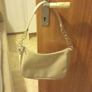  Axelväska med dragkedja - Säljer en snygg axelväska i syntetmaterial. Väskan har en praktisk dragkedja och en stilren design med silverfärgade kedjedetaljer på axelremmen. Perfekt för vardagsbruk eller en kväll ute. Passar till alla säsonger och outfits! Den är oanvänd. 