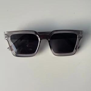 Grå rektangulära solglasögon - Snygga rektangulära solglasögon med transparent grå båge och mörka linser. Perfekta för en stilren look, oavsett om du är på stranden eller i stan. De har en modern design som passar till alla outfits. Ett måste för soliga dagar!