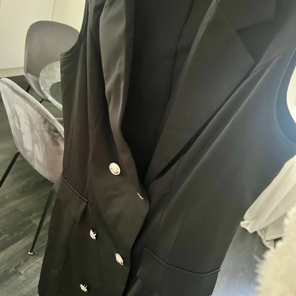 Snygg svart ärmlös klänning med dubbelknäppning och stiliga knappar. Perfekt för en elegant look, kanske till en fest eller ett speciellt tillfälle. Klänningen har en klassisk krage och är i en stilren design.. Mekot.