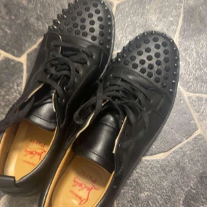 Svarta sneakers med nitar - Säljer ett par svarta sneakers från Christian Louboutin i skinn med coola nitar på tån. De har den klassiska röda sulan och snörning framtill. Perfekta för att ge din outfit en edgy touch! Använda men i bra skick.