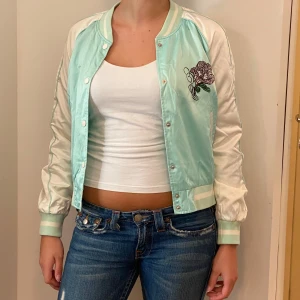 Mintgrön bomberjacka med broderi - Säljer en superfin mintgrön bomberjacka från H&M. Den har ett snyggt blommigt broderi på bröstet och ryggen. Jackan är i satinmaterial och har långa ärmar med ribbade muddar. Perfekt för vår och sommar! 🌸