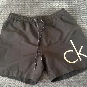 Säljer ett par snygga svarta badshorts från Calvin Klein i storlek S. De har en elastisk midja med dragsko och en praktisk bakficka med dragkedja. Perfekta för sommarens strandhäng eller poolpartyn! 🏖️