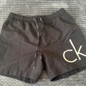 Svarta badshorts från Calvin Klein - Säljer ett par snygga svarta badshorts från Calvin Klein i storlek S. De har en elastisk midja med dragsko och en praktisk bakficka med dragkedja. Perfekta för sommarens strandhäng eller poolpartyn! 🏖️