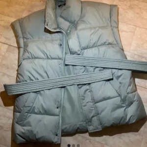 Grå dunväst med bälte - Säljer en snygg grå dunväst med bälte. Perfekt för höst och vinter! Västen har en hög krage och knappar framtill. Den är både stilren och praktisk, idealisk för lager-på-lager under kyliga dagar. Passar bra till både vardag och lite finare tillfällen.