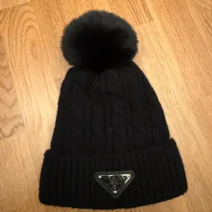 Snygg svart stickad mössa från dhgate med en stor fluffig pälsboll på toppen. Mössan har ett ribbat uppvik och en metallplatta med logotyp framtill. Perfekt för att hålla sig varm och stilren under kalla dagar.