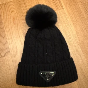 Svart stickad mössa med pälsboll - Snygg svart stickad mössa från dhgate med en stor fluffig pälsboll på toppen. Mössan har ett ribbat uppvik och en metallplatta med logotyp framtill. Perfekt för att hålla sig varm och stilren under kalla dagar.