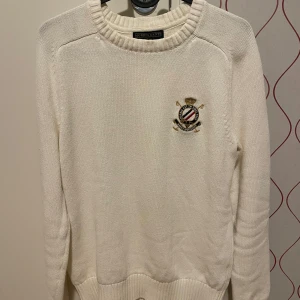 Vit stickad tröja från Morris Gents - Säljer en stilren vit stickad tröja från Morris Gents i storlek XS. Tröjan har ett broderat emblem på bröstet och ribbade muddar vid ärmslut och nederkant. Perfekt för en klassisk och elegant look. Passar både till vardags och festliga tillfällen.