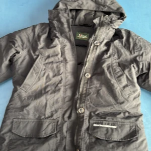 Svart pufferjacka från Peak Performance - Säljer en svart pufferjacka från Peak Performance. Den är perfekt för kalla dagar med sin vadderade insida och huva. Jackan har dragkedja och knappar framtill samt praktiska fickor. Perfekt för vintern och hösten!