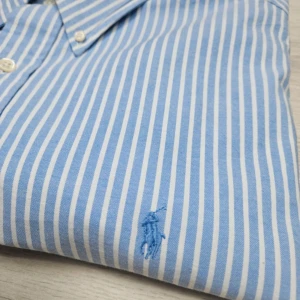 Ralph Lauren Blå och vit randig skjorta - Säljer en klassisk blå och vit randig skjorta från Ralph Lauren. Skjortan är långärmad och har en broderad logga på bröstet. Perfekt för både vardag och fest! Den är i bomull och har en normal passform med knappar framtill.