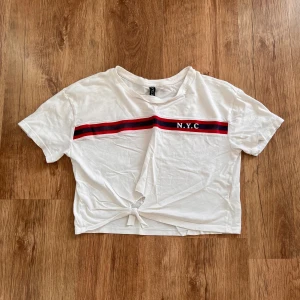 Vit kortärmad topp från H&M med NYC-tryck - Säljer en vit kortärmad topp från H&M i storlek XS. Den har ett coolt NYC-tryck med en röd och svart rand över bröstet. Toppen är croppad och har en knytning framtill för en avslappnad look. Perfekt för en casual dag ute!