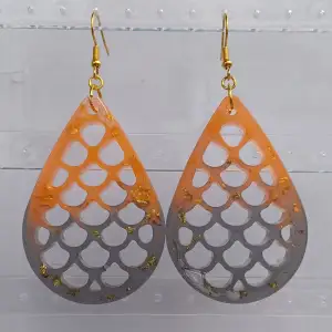 Snygga droppformade örhängen i en cool orange och svart färgkombination. De har ett unikt mönster med gulddetaljer som ger en extra touch av glamour. Perfekta för att liva upp vilken outfit som helst! Gjorda i resin. OBS bilden är ett exempel, viss färg/glitter skillnad kan förekomma.