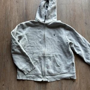 Grå hoodie från Polo Ralph Lauren - Säljer en grå hoodie från Polo Ralph Lauren i storlek L. Barn 14-16år Den har en dragkedja framtill och en klassisk känguruficka. Perfekt för en avslappnad stil och passar bra till både höst och vår. Hoodien är i bra skick och superbekväm!