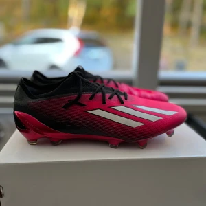 Rosa och svarta fotbollsskor från Adidas - Säljer ett par snygga fotbollsskor från Adidas i rosa och svart. Skorna har en modern design med tre vita ränder på sidan och svarta snören. De är perfekta för fotbollsträning och matcher. Sulan har flera dobbar för bra grepp på planen.