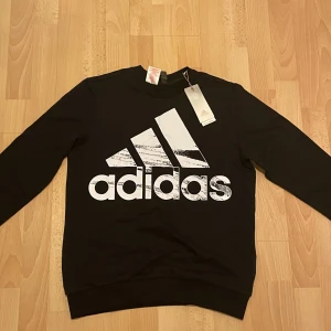 Svart sweatshirt från Adidas - Säljer en snygg svart sweatshirt från Adidas i nyskick. Den har en stor vit Adidas-logga på framsidan och är perfekt för både vardag och träning. Tröjan har långa ärmar och är superbekväm. Passar perfekt till höst och vinter! Helt ny aldrig använd!