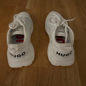 Vita sneakers från Hugo - Säljer ett par snygga vita sneakers från Hugo Boss. De har en stilren design med snörning och märkets logga tydligt synlig på hälen. Perfekta för en casual look och passar till det mesta i garderoben. Använda max 5 ggr men i superbra skick!