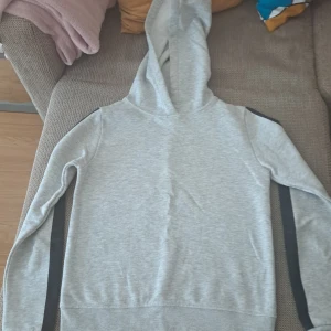 Grå hoodie med svarta detaljer - Säljer en stilren grå hoodie med svarta ränder längs ärmarna. Perfekt för en avslappnad look. Den är supermjuk och bekväm, perfekt för både höst och vår. Passar bra till jeans eller joggers!