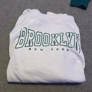 Vit sweatshirt med Brooklyn-tryck - Säljer en vit sweatshirt med texten 'Brooklyn New York' i grönt på framsidan. Tröjan har långa ärmar och en avslappnad passform, perfekt för en casual look.