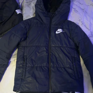 Svart dunjacka från Nike - Säljer en svart dunjacka från Nike med huva och dragkedja. Jackan har en klassisk design med Nike-logga på bröstet. Perfekt för kyliga dagar.
