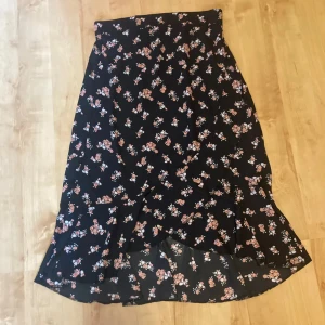 Svart blommig kjol - Snygg svart kjol med blommigt mönster i rosa och beige toner. Kjolen har en volangdetalj längst ner som ger en fin rörelse. Perfekt för både vardag och speciella tillfällen.