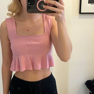 Rosa ribbad topp med volang - Säljer en söt rosa ribbad topp med volangdetalj i nederkant. Toppen är ärmlös och har en fyrkantig urringning. Perfekt för sommarens varma dagar!